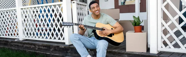 Счастливый африканский американец, играющий на акустической гитаре возле коробок на крыльце нового дома, недвижимости, баннера — стоковое фото