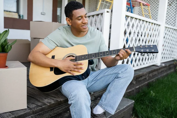 Веселый африканский американец играет на акустической гитаре рядом с картонными коробками и новым домом — стоковое фото