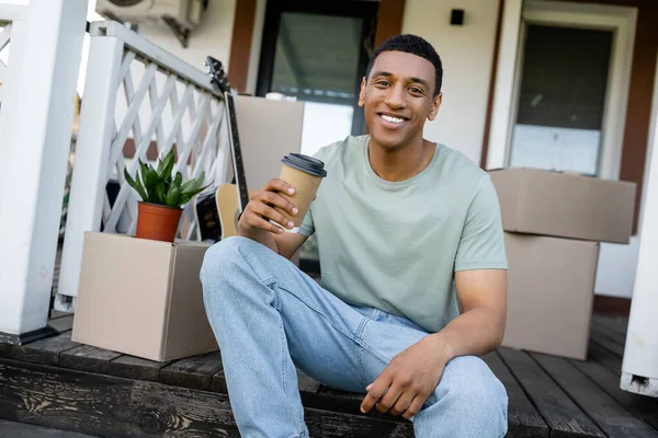 Fröhlicher afrikanisch-amerikanischer Mann hält Kaffee in der Nähe von Kartons und neuem Haus im Freien — Stockfoto