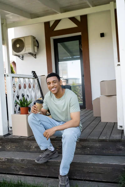 Sorrindo afro-americano homem segurando café para ir perto de pacotes no alpendre da nova casa — Fotografia de Stock