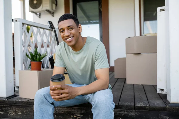 Веселый африканский американец держит кофе на вынос рядом с коробками на крыльце нового дома — стоковое фото