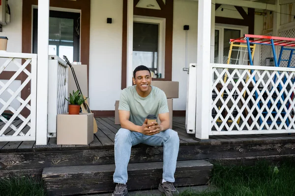 Усміхнений афроамериканський чоловік тримає каву на винос, сидячи на ганку нового будинку — стокове фото