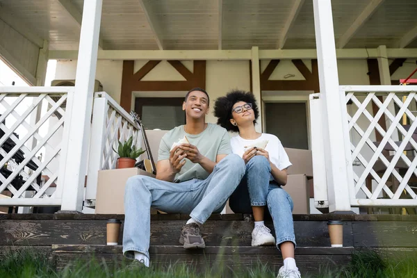 Positive afrikanisch-amerikanische Paar hält Sandwiches in der Nähe von Kaffee auf der Veranda des neuen Hauses gehen — Stockfoto