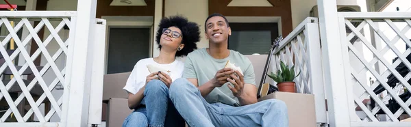 Задоволена афроамериканська пара тримає смачні бутерброди на ганку нового будинку, нерухомості, банера — стокове фото