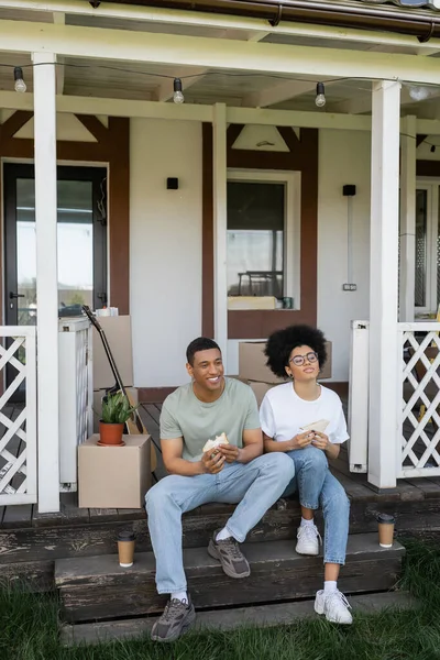 Joyeux couple afro-américain tenant des sandwichs près du café et des paquets sur le porche de la nouvelle maison — Photo de stock