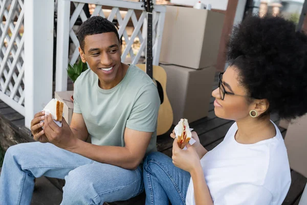 Souriant homme afro-américain tenant sandwich près de petite amie et des boîtes sur le porche de la nouvelle maison — Photo de stock