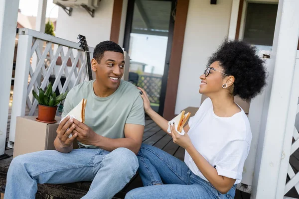 Allegra coppia afro-americana parlando e tenendo panini sul portico della nuova casa — Foto stock