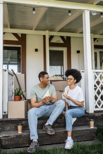 Feliz afroamericana mujer celebración sándwich cerca de novio y café para ir en porche de nueva casa - foto de stock