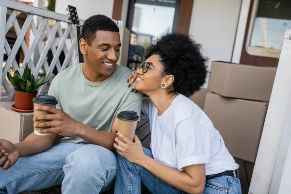 Улыбающаяся африканская американская пара держит кофе на вынос и разговаривает на крыльце нового дома — стоковое фото