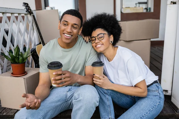 Sourire couple afro-américain avec café pour aller parler près des boîtes sur le porche de la nouvelle maison — Photo de stock