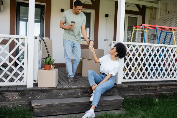 Позитивный африканский американец дает кофе на вынос подруге возле коробок и нового дома — стоковое фото