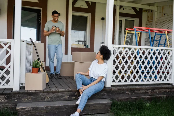 Felice uomo africano americano in possesso di caffè per andare vicino fidanzata e scatole sul portico della nuova casa — Foto stock