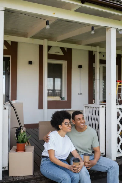 Lächelnd afrikanisch-amerikanischer Mann umarmt Freundin mit Kaffee in der Nähe von Paketen auf der Veranda des neuen Hauses — Stockfoto