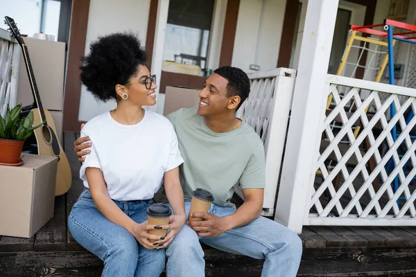 Позитивная африканская американская пара обнимает и держит кофе, чтобы подойти к коробкам на крыльце нового дома — стоковое фото