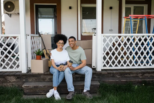 Улыбающаяся африканская американская пара держит кофе, чтобы подойти к пакетам на крыльце нового дома — стоковое фото