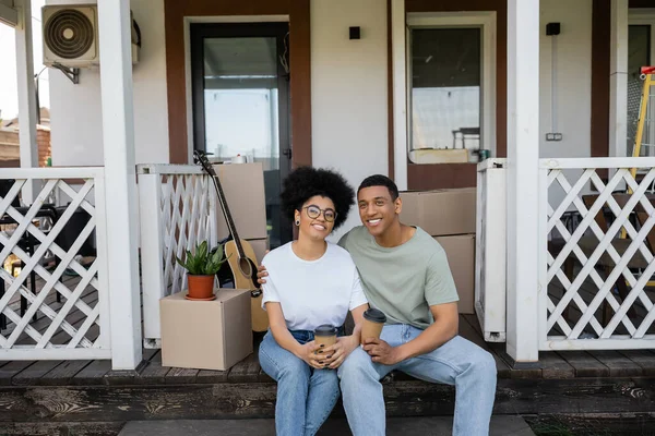 Счастливая африканская американская пара держит кофе на вынос и обнимается на крыльце нового дома — стоковое фото