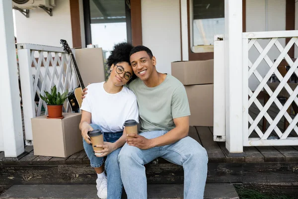 Smiling african american couple with coffee to go hugging while sitting on couch of new house — Stock Photo