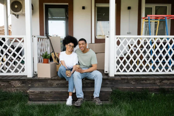 Fröhliche afrikanisch-amerikanische Paar umarmt und hält Kaffee auf der Veranda des neuen Hauses gehen — Stockfoto
