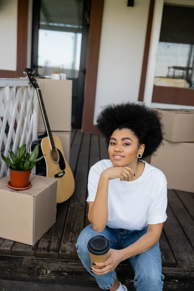 Positif afro-américaine femme tenant café pour aller et regardant caméra près de nouvelle maison — Photo de stock