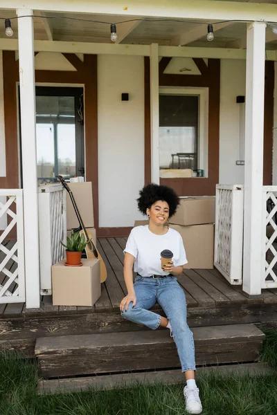 Femme afro-américaine joyeuse avec du café pour aller assis sur le porche près des boîtes et de la nouvelle maison — Photo de stock
