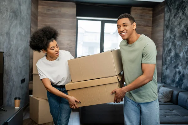 Positivo Africano americano casal segurando caixas de papelão durante a mudança na sala de estar — Fotografia de Stock