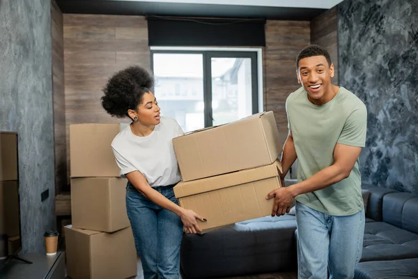 Cheerful african american man holding packages with girlfriend in living room in new house — Stock Photo