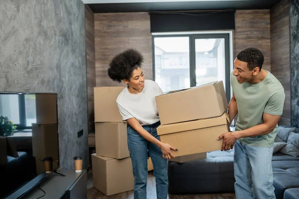 Pareja afroamericana positiva sosteniendo cajas de cartón y sonriendo en la sala de estar en casa nueva - foto de stock