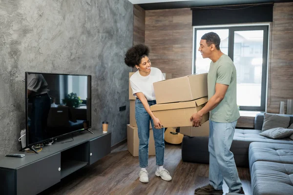 Alegre casal afro-americano segurando caixas de papelão perto de sofá e tv na nova sala de estar — Fotografia de Stock