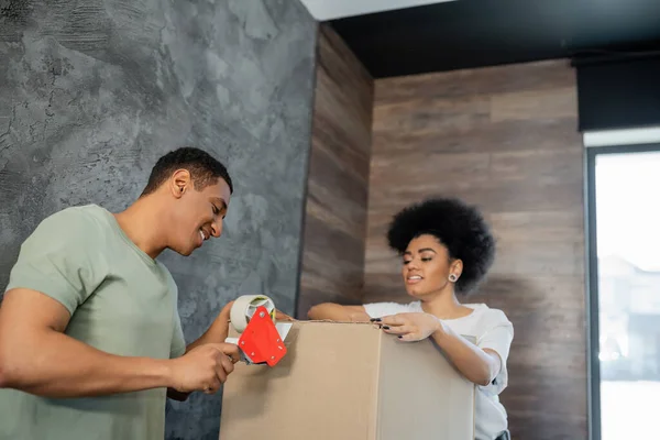 Smiling african american man holding adhesive tape near cardboard boxes and girlfriend during moving — Stock Photo