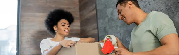 Усміхнений афроамериканський чоловік тримає клейку стрічку біля коробки і дівчину під час руху, банер — стокове фото