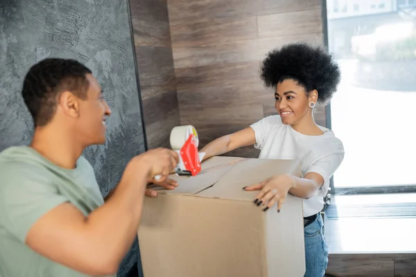 Улыбающаяся африканская американка держит коробку рядом с парнем с клейкой лентой во время переезда — стоковое фото