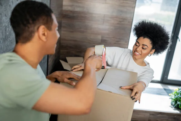 Positive african american couple packaging cardboard box with adhesive tape during relocation — Stock Photo