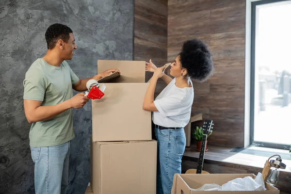 Side view of happy african american couple with adhesive tape packaging boxes during moving — Stock Photo