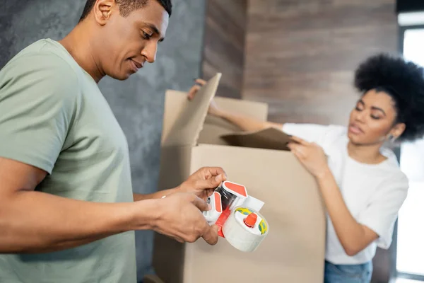 African american man holding adhesive tape near blurred girlfriend with boxes during moving — Stock Photo