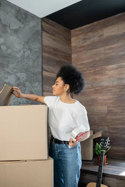 Side view of african american woman holding adhesive tape near carton boxes during moving — Stock Photo