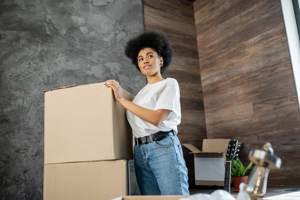 African american woman standing near cardboard boxes in living room in new house — Stock Photo