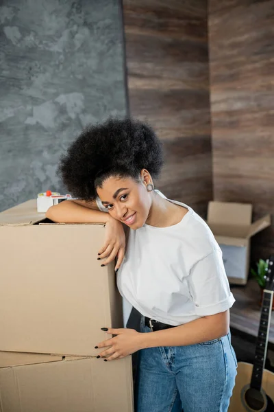 Cheerful african american woman standing near carton boxes and adhesive tape in living room — Stock Photo