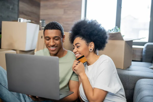 Coppia afro-americana positiva utilizzando laptop e carta di credito durante il trasferimento in una nuova casa — Foto stock