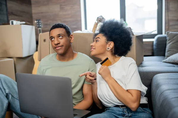 Afro-américaine détenant une carte de crédit et pointant vers un ordinateur portable près de petit ami lors de la relocalisation — Photo de stock