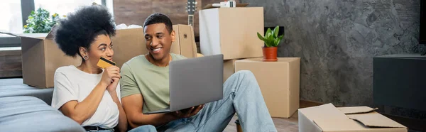 Позитивна афроамериканська пара використовує ноутбук і кредитну картку під час переїзду в новому будинку, банер — стокове фото