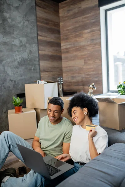 Positif couple afro-américain faisant des achats en ligne près de boîtes en carton dans une nouvelle maison — Photo de stock