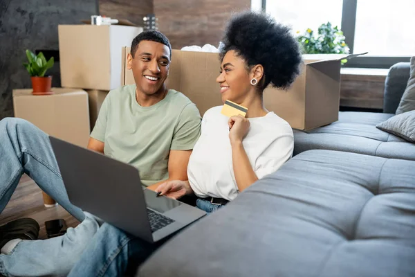 Улыбающаяся африканская американка, пользующаяся ноутбуком и кредитной картой рядом с парнем и коробками в новом доме — стоковое фото
