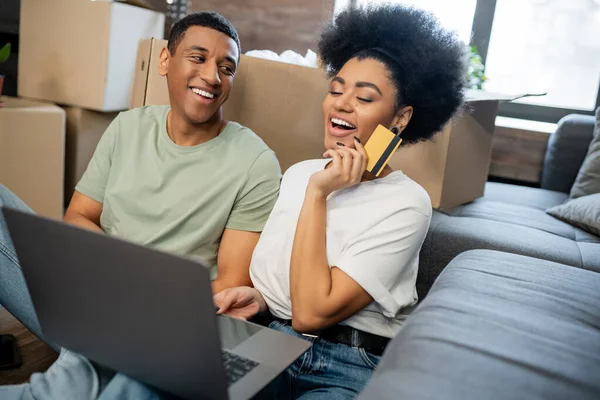 Excited african american woman doing online shopping near boyfriend and carton boxes in new home — Stock Photo