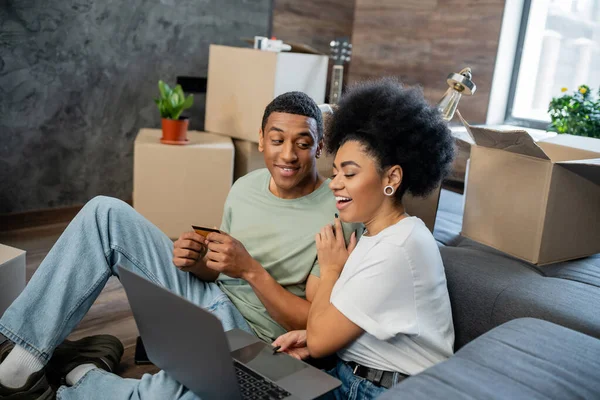 Despreocupado Africano americano mulher abraçando namorado com laptop e cartão de crédito perto de caixas em casa — Fotografia de Stock