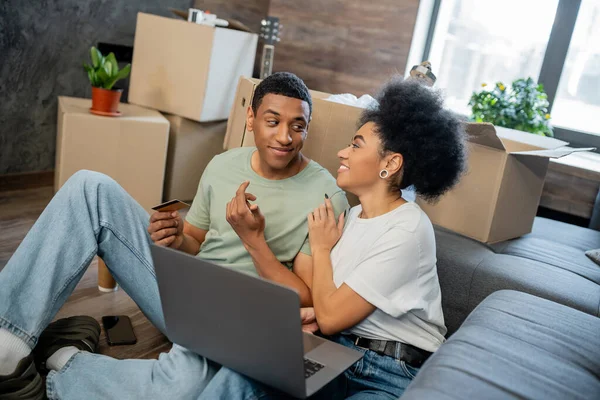 Joyeuse femme afro-américaine parlant à son petit ami lors des achats en ligne près de boîtes dans une nouvelle maison — Photo de stock