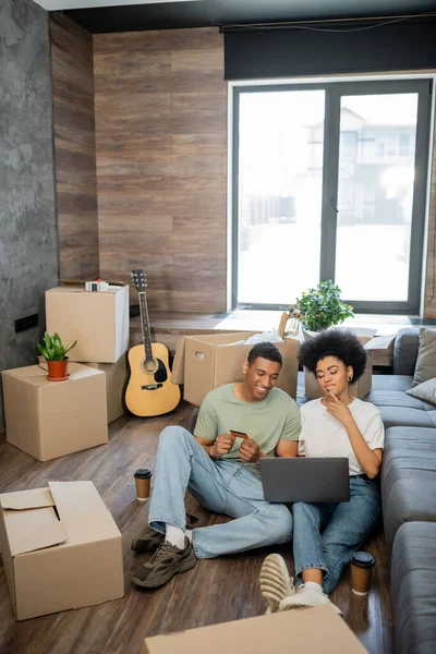 Позитивная африканская американская пара делает онлайн покупки возле кофе и коробки в новом доме — стоковое фото