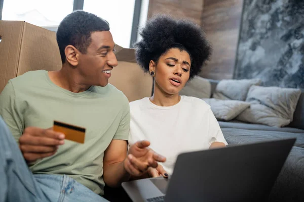 Chateado afro-americano mulher usando laptop perto sorrindo namorado e caixas de papelão em nova casa — Fotografia de Stock