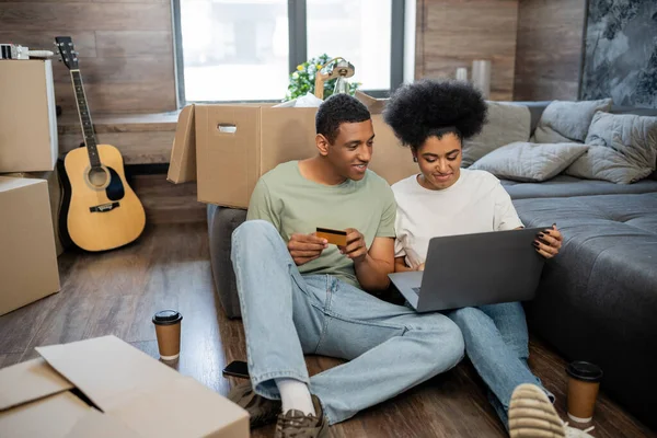 Homme afro-américain tenant carte de crédit près de petite amie avec ordinateur portable et boîtes dans une nouvelle maison — Photo de stock
