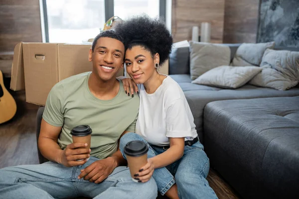 Улыбающаяся африканская американская пара держит кофе, чтобы подойти к коробке в новой гостиной — стоковое фото