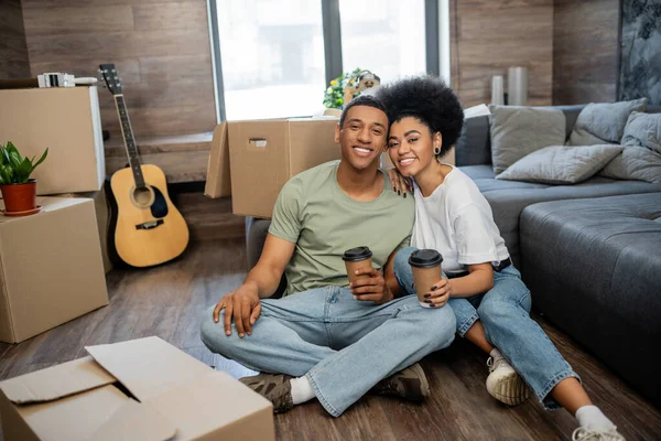 Heureux couple afro-américain avec café regardant la caméra près des paquets dans le nouveau salon — Photo de stock